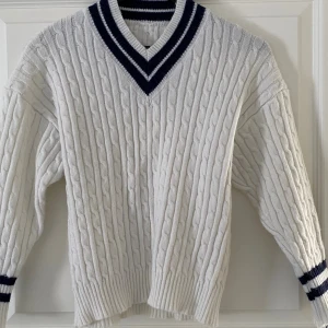 Vit kabelstickad tröja från SHEIN - Kabelstickad vit tröja från SHEIN med mörkblå ränder vid v-ringad krage och ärmslut. Tröjan har lång ärm, ribbade muddar och klassisk preppy stil. Perfekt för dig som gillar sportig och stilren look.