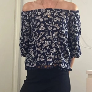 Mörkblå offshoulder blus med mönster - Snygg mörkblå offshoulder blus med vitt paisleymönster och trekvartsärmar. Blusen har en lös passform och dekorativa bollar längs nederkanten. Perfekt för dig som gillar boho-stil och vill ha något luftigt och bekvämt.