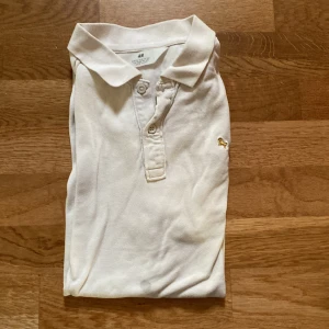 Vit pikétröja från H&M - Klassisk vit pikétröja från H&M med krage och knappar framtill. Tröjan har korta ärmar och en liten broderad detalj på bröstet. Perfekt för en clean och stilren look. Tillverkad i mjuk bomull som känns skön mot huden. Det finns en liten fläck men den syns knappt (går att se på första bilden längst ner på tröjan)