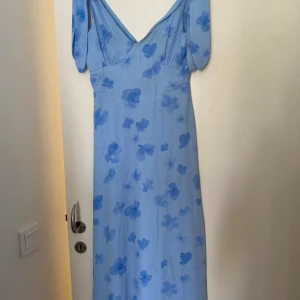 Ljusblå långklänning med blommor - Superfin ljusblå långklänning med blommigt mönster och v-ringad hals. Klänningen har korta puffärmar och en smickrande passform. Perfekt för sommarens alla tillfällen och ger en romantisk vibe.