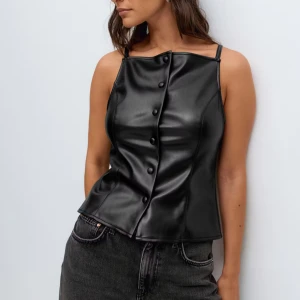 Svart skinnkorsett med knappar xs - Oanvänd snygg svart korsettopp i skinnimitation med smala axelband och dekorativa knappar framtill. Toppen har en figurnära passform och en stilren, modern look som passar perfekt till jeans eller kjol. Från Gina i Stl xs. 