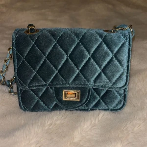 Quiltad blå axelväska. - Supersnygg quiltad axelväska i blå sammet med guldfärgad kedja och vridlås. Väskan har ett klassiskt rutmönster och en lyxig känsla med mjuk yta. Från Ur och Penn.