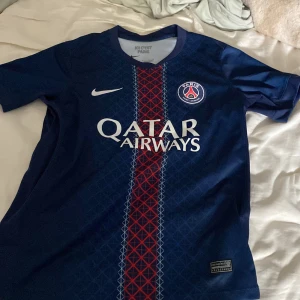 PSG matchtröja Nike Dri-FIT blå S - Paris Saint-Germain fotbollströja från Nike i Dri-FIT-material. Djupblå med röd och vit rand i mitten, PSG-logga på bröstet och Qatar Airways-tryck. Kortärmad och med geometriskt mönster framtill. Perfekt för dig som älskar fotboll och vill sticka ut.