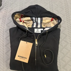 Svart hoodie från Burberry med dragkedja - Svart hoodie från Burberry med klassiskt rutigt foder i huvan, guldfärgade detaljer och dragkedja framtill. Tröjan har snörning i huvan och känguruficka. Perfekt för dig som vill ha en stilren och lyxig streetwear-look. Dessutom helt ny och aldrig använd, köpte fel storlek