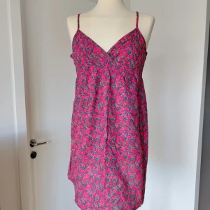 Rosa blommig sommarklänning med smala axelband - Söt kort klänning med smala axelband och v-ringning. Klänningen är rosa med blommigt mönster, har smock över bysten och är tillverkad i ett lätt och luftigt material. Perfekt för varma dagar och ger en avslappnad vibe.