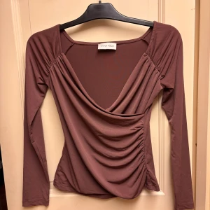 Brun omlottopp från Anna Field - Säljer en brun omlottopp från Anna Field med lång ärm och snygg drapering framtill. Toppen har v-ringning och rynkade detaljer i sidan. Står ingen storlek men sitter som en XS!
