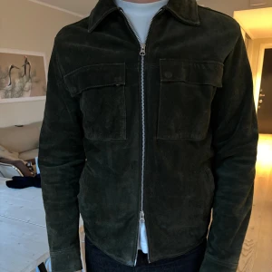 Olivgrön mockajacka från Human Scales - Snygg olivgrön mockajacka från Human Scales i storlek S. Jackan har en dragkedja framtill och två stora bröstfickor. Tillverkad av 100% koskinn med polyesterfoder. Perfekt för höst och vår! Tecken på användning finns.