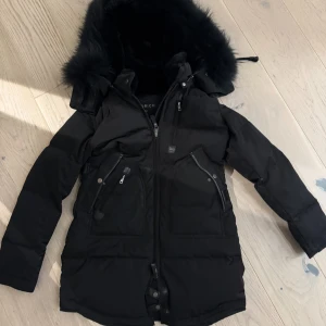 Svart dunjacka Cedrico  - Svart dunjacka med stor huva , flera dragkedjeförsedda fickor och snygga detaljer. Jackan är lång, har hög krage och är perfekt för kalla dagar. Klassisk och stilren design som håller dig varm hela vintern. Storlek xs rak modell 