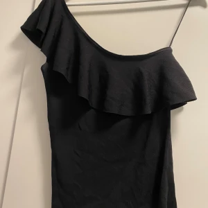 Svart oneshoulder topp med volang - Säljer en svart oneshoulder topp med snygg volangdetalj längs halsringningen. Toppen är croppad och har en tight passform som framhäver figuren.🖤💘