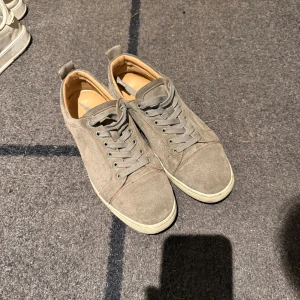Christian Louboutin sneakers mocka grå - Säljer ett par grå sneakers från Christian Louboutin i mjuk mocka med klassisk röd sula och snörning. Skorna har rund tå, beige innersida i skinn och diskreta detaljer. Perfekta för dig som vill ha en clean och lyxig look. Skorna är smutsiga men i bra skick, har inte låda osv. Hör av er om ni har mer frågor!