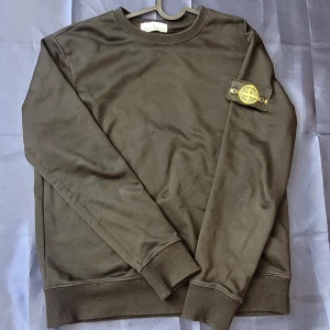 Svart sweatshirt från Stone Island - Svart sweatshirt från Stone Island med klassisk rund hals och ribbade muddar. Tröjan har den ikoniska Stone Island-loggan på vänster ärm. Tillverkad i mjuk bomull för skön komfort och stilren look. Perfekt för dig som gillar streetwear och snygga detaljer.
