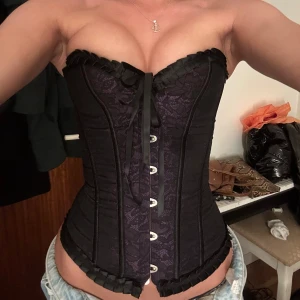 Svart och lila korsett med spetsdetaljer - Säljer en snygg korsett i svart och lila med spetsmönster och dekorativ snörning bak. Toppen är axelbandslös och har volangkant upptill samt knäppning framtill. Perfekt för dig som vill ha en edgy och feminin look.