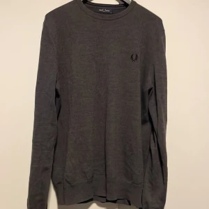 Mörkgrå tröja från Fred Perry - Snygg mörkgrå tröja från Fred Perry med diskret logga broderad på bröstet. Tröjan har rund hals, långa ärmar och ribbade muddar. Perfekt för dig som gillar stilrena och klassiska plagg med en sportig touch.