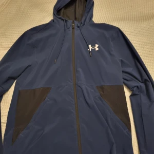Blå jacka från Under Armour - Mörkblå vindjacka från Under Armour med svarta detaljer på sidorna och vitt logotryck på bröstet. Jackan har huva med snörning, hel dragkedja och meshfoder på insidan. 