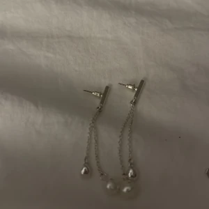 Silverfärgade örhängen med kedjor och pärlor - Säljer ett par stilrena örhängen i silverfärg med två tunna kedjor som hänger ner från en minimalistisk stav. I änden av kedjorna sitter en liten silverkula och en vit pärla som ger en snygg och modern look.