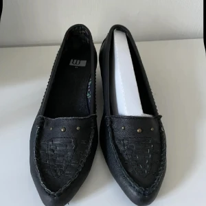 Svarta skinn  loafers med liten klack 37 nya  - Stilrena svarta loafers med spetsig tå och diskret liten klack. Skorna har dekorativa nitar framtill och flätad detalj på ovandelen. Tillverkade i syntetmaterial med synliga sömmar för en klassisk look.