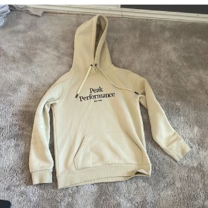 Beige hoodie från Peak Performance - Snygg beige hoodie från Peak Performance med svart logotyp på bröstet. Tröjan har huva med snörning, magficka och ribbade muddar vid ärmslut och nederkant. Perfekt för chill dagar och enkel att matcha med jeans eller joggers.