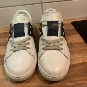 Valentino opens  - Snygga vita Valentino opens sneakers med bred svart rem över vristen och ikoniska nitar runt hälen. Skorna har snörning som kan has i skon. Stil rena skor som passar bra nu till vintern