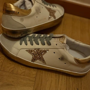 Golden Goose sneakers med glitterstjärna - Säljer ett par Golden Goose sneakers i vitt skinn med grå mockadetaljer och grå snörning. Skorna har en glittrig guldstjärna på sidan och gulddetaljer på hälen samt guldtext på plösen. Sulan har en sliten look och insidan är vit. Vet inte om dom e äkta fick dom 