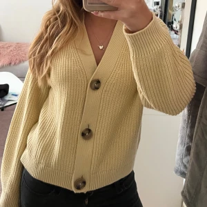 Beige kofta från H&M med stora knappar - Superfin beige kofta från H&M i stickad bomull. Koftan har stora bruna knappar framtill, v-ringning och ribbade muddar. Perfekt att slänga över en topp för en chill och snygg look. Passar dig som gillar en lite loose fit.