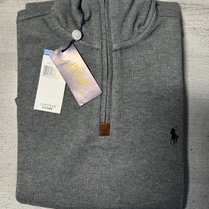Grå half zip tröja från Polo Ralph Lauren - Snygg grå tröja från Polo Ralph Lauren med half zip och klassisk broderad logga på bröstet. Tröjan har hög krage, långa ärmar och är tillverkad i mjuk bomullsmix. Perfekt för lager-på-lager och stilren look. TRÖJAN är aldrig använd! Köpte på kryssnings resa för 125$.