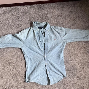 Ljusblå skjorta från Polo Ralph Lauren - Klassisk ljusblå skjorta från Polo Ralph Lauren med broderad logga på bröstet. Skjortan har knappar hela vägen, button-down-krage och långa ärmar. Skicket är även som nytt!