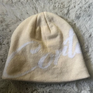 Beige stickad mössa från Corteiz - Snygg beige mössa från Corteiz med vit, stor logotyp i skriven stil över hela mössan. 