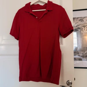 Röd pikétröja från Dressmann L - Röd pikétröja från Dressmann i storlek L. Klassisk krage, kort ärm och två knappar vid halsen. Tillverkad i mjuk bomull som känns skön mot huden. Enkel och stilren design utan mönster eller tryck.