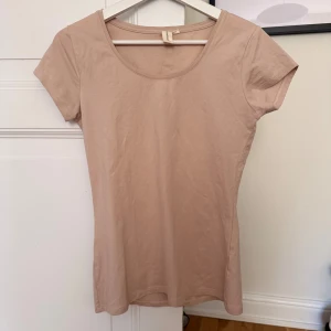 Beige-ljusrosa basic t-shirt från SunShine - Enkel och stilren basic beige t-shirt från Sunshine i mjuk bomull. Modellen är figurnära med rund halsringning och korta ärmar. Perfekt att matcha med jeans eller kjol för en clean look.