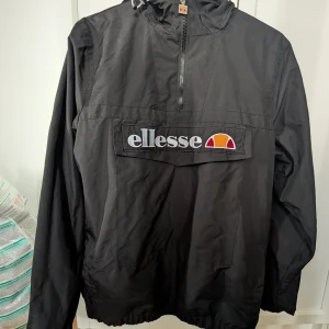 Svart vindjacka från Ellesse - Svart vindjacka från Ellesse med stor logga framtill och patch på ärmen. Jackan har huva, half zip-dragkedja och elastiska muddar. Tillverkad i polyester och har en sportig, loose passform. Perfekt för dig som gillar streetwear och snygga detaljer. OBS. Är sydd i ena armhålan. 