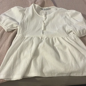 Vit blus med knyt fram från 157 - Supersöt vit blus från 157 med korta puffärmar och två knyt fram. Blusen har en lätt strukturerad yta och en lös, flowy passform med markerad midja. Perfekt till jeans eller kjol för en fräsch look.