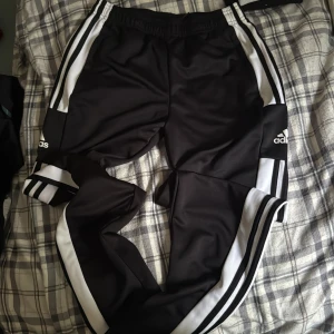 Svarta Adidas träningsbyxor med ränder - Svarta träningsbyxor från Adidas med klassiska vita ränder längs sidorna och Adidas-logga på låret. Byxorna har resår i midjan och är tillverkade i ett glansigt syntetmaterial. Perfekta för sport eller chill, med en loose fit och sportig look.