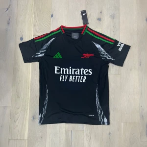 Svart Arsenal fotbollströja Adidas - Snygg svart Arsenal fotbollströja från Adidas med gröna och röda detaljer på axlarna, klubbmärke i rött och vit Emirates-sponsor på bröstet. Tröjan har korta ärmar och är tillverkad i ett lätt och ventilerande material.