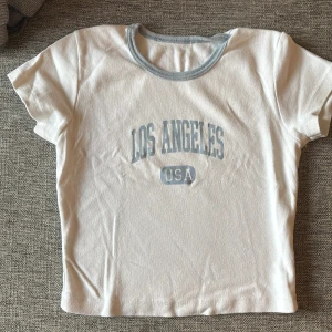 Vit t-shirt med Los Angeles-tryck - Vit t-shirt med ljusblå text 'Los Angeles USA' på bröstet och matchande ljusblå kant vid halsen. Klassisk kortärmad modell i mjuk bomull, perfekt för en avslappnad och trendig look.