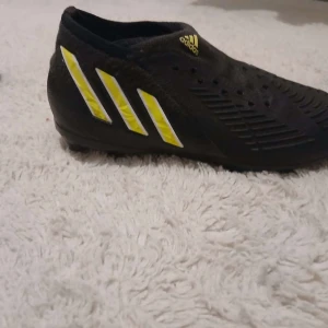 Adidas Predator svarta fotbollsskor - Säljer ett par svarta Adidas Predator fotbollsskor med gula och vita detaljer. Skorna har klassiska tre ränder på sidan, låg profil och mönstrad ovansida för extra grepp. Yttersulan har fasta dobbar i orange och svart. Perfekta för dig som vill dominera på planen.
