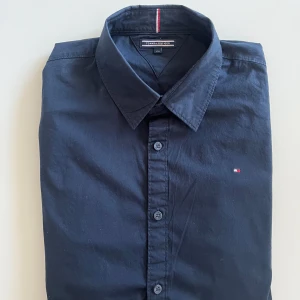 Mörkblå skjorta från Tommy Hilfiger - Snygg mörkblå skjorta från Tommy Hilfiger med klassisk krage och knappar hela vägen. Liten broderad logga på bröstet och långa ärmar. Skjortan är i mjuk bomull och passar perfekt till en stilren look. Storlek S