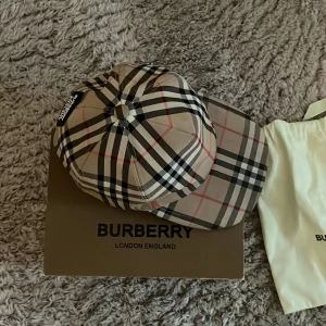 Burberry rutig keps i beige och svart - Säljer en klassisk Burberry keps med det ikoniska rutmönstret i beige, svart, vitt och röda detaljer. Kepsen har justerbar rem bak och en patch med Burberry London England. Materialet känns slätt och exklusivt, perfekt för dig som gillar stilrena accessoarer.