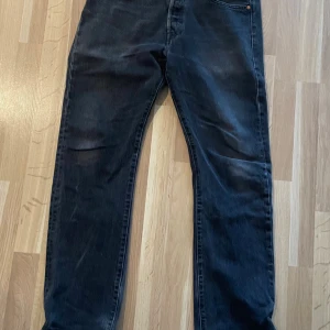 Levi's 501 svarta jeans W34 L34 - Svarta Levi's 501 jeans med klassisk rak passform och knappgylf. Jeansen är tillverkade i slitstarkt denim och har en mörk tvätt med lätt slitning på vissa partier. Perfekta för dig som gillar en tidlös och avslappnad stil.