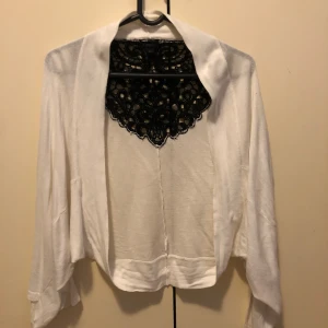 Vit kofta med svart spetsdetalj - Säljer en vit kofta från KINDLY med lång ärm och snygg svart spetsdetalj vid halsen. Koftan har en öppen front och är tillverkad i ett mjukt material som känns skönt mot huden. Perfekt för lager-på-lager och ger en cool kontrast med spetsen.