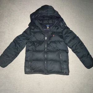 Svart pufferjacka från Polo Ralph Lauren - Svart pufferjacka från Polo Ralph Lauren med huva och dragkedja framtill. Jackan har det klassiska röda Polo-logot broderat på bröstet och två sidofickor med dragkedja. Perfekt för kalla dagar och har en snygg, stilren look. Barnstorlek men sitter som s