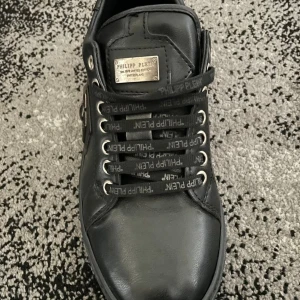 Svarta sneakers från Philipp Plein - Snygga svarta sneakers från Philipp Plein i skinn med silverdetaljer och logotyp på sidan. Skon har snörning med märkta skosnören och metallplatta på plösen. Sulan pryds av Philipp Plein-text i silver. Perfekt för dig som gillar exklusiv streetstyle.