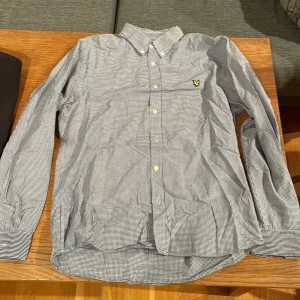 Rutig skjorta från Lyle & Scott, blå/vit - Snygg rutig skjorta från Lyle & Scott i blått och vitt med klassisk button down-krage och broderad gul logga på bröstet. Skjortan är långärmad och har vita knappar framtill. Perfekt för dig som gillar stilren och tidlös design.