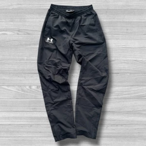 Svarta träningsbyxor från Under Armour - Svarta träningsbyxor från Under Armour med elastisk midja och dragsko. Byxorna har en loose passform och är tillverkade i lätt syntetmaterial med meshfoder för extra komfort. Perfekta för dig som vill ha bekväma och funktionella byxor till träning.