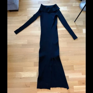 Svart långärmad offshoulder klänning MNG - Svart långklänning från Mango med offshoulder-krage och lång ärm. Klänningen är ribbad, har en slits framtill och är figurnära i modellen. Tillverkad i en mix av polyester och polyamid för en stretchig och bekväm känsla.