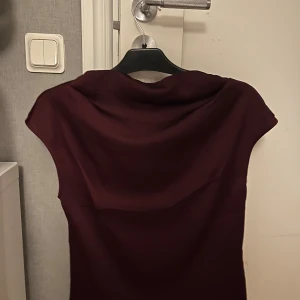 Vinröd blus med drapering - Säljer en vinröd blus med snygg draperad halsringning och korta ärmar. Blusen har en stilren och enkel design som ger ett sofistikerat intryck. Perfekt för dig som gillar minimalistisk stil och vill sticka ut med en djup färg. Storlek XS, aldrig använd❣️ märket är HM