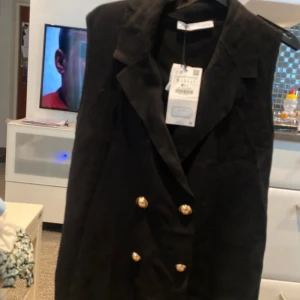 Svart lång väst från Zara med guldknappar - Snygg svart lång väst från Zara med klassisk krage och dubbelknäppning med fyra guldiga knappar framtill. Västen har en rak passform och är tillverkad i ett mjukt polyestermaterial. Perfekt att styla över en t-shirt eller skjorta för en cool look.