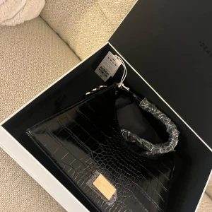 Svart axelväska från IDEAL OF SWEDEN - Nypris 1990kr. Snygg svart axelväska med croco-mönster från IDEAL OF SWEDEN. Väskan har en stilren rektangulär form, guldfärgade detaljer och en bred axelrem. Perfekt för dig som vill ha en trendig accessoar med lyxig känsla. Materialet är glansigt syntet med croco-textur. Justerbarband.