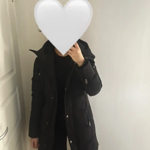 Svart lång dunjacka Rockandblue - Svart lång dunjacka från Rockandblue med huva och snygga detaljer som dragkedjor och knappar framtill. Jackan har en stilren look, emblem på ärmen och är perfekt för kalla dagar. Flera fickor med dragkedja och quiltad design för extra värme.