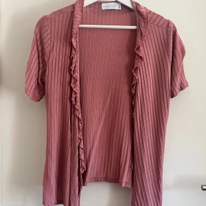 Rosa ribbad blus med volang  - Säljer en söt rosa blus från Three Sixty Five i storlek small. Blusen har korta ärmar, ribbat tyg och volangdetaljer längs den öppna framkanten. 