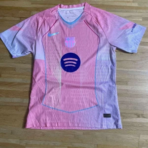 Barça x Nike rosa fotbollströja - Säljer en Barcelona fotbollströja i rosa och ljuslila med Nike-logga, klubbmärke och Spotify-tryck på bröstet. UNHCR-logga på ryggen. Tröjan har korta ärmar och är tillverkad i ett lätt, ventilerande material perfekt för match eller träning.